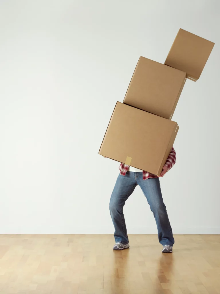 Personne portant une pile de trois cartons de déménagement empilés, vêtue d'une chemise à carreaux rouge et blanc et d'un